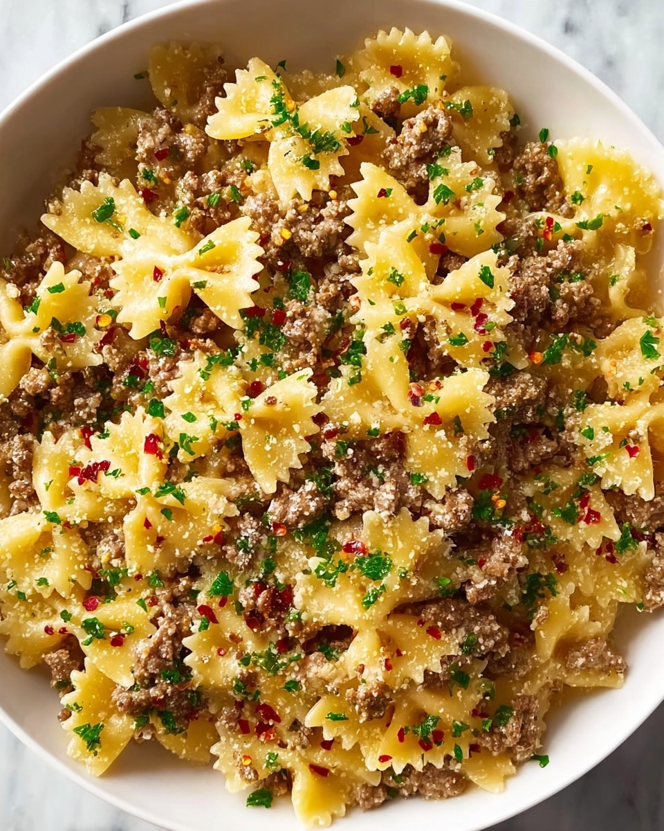 The image shows a white bowl filled with farfalle pasta coated in a light sauce, mixed evenly with ground meat scattered throughout. The pasta is a warm yellow color with a slightly glossy texture, and the ground meat is brown, crumbly, and spread over the top and below the pasta layers. Small chopped green herbs are sprinkled generously over the dish, adding bright green spots, and tiny red chili flakes are scattered for a touch of color and spice. The dish has a light dusting of grated cheese that adds a slightly grainy texture on top. The bowl sits on a white marbled surface. photo taken with an iphone --ar 4:5 --v 7