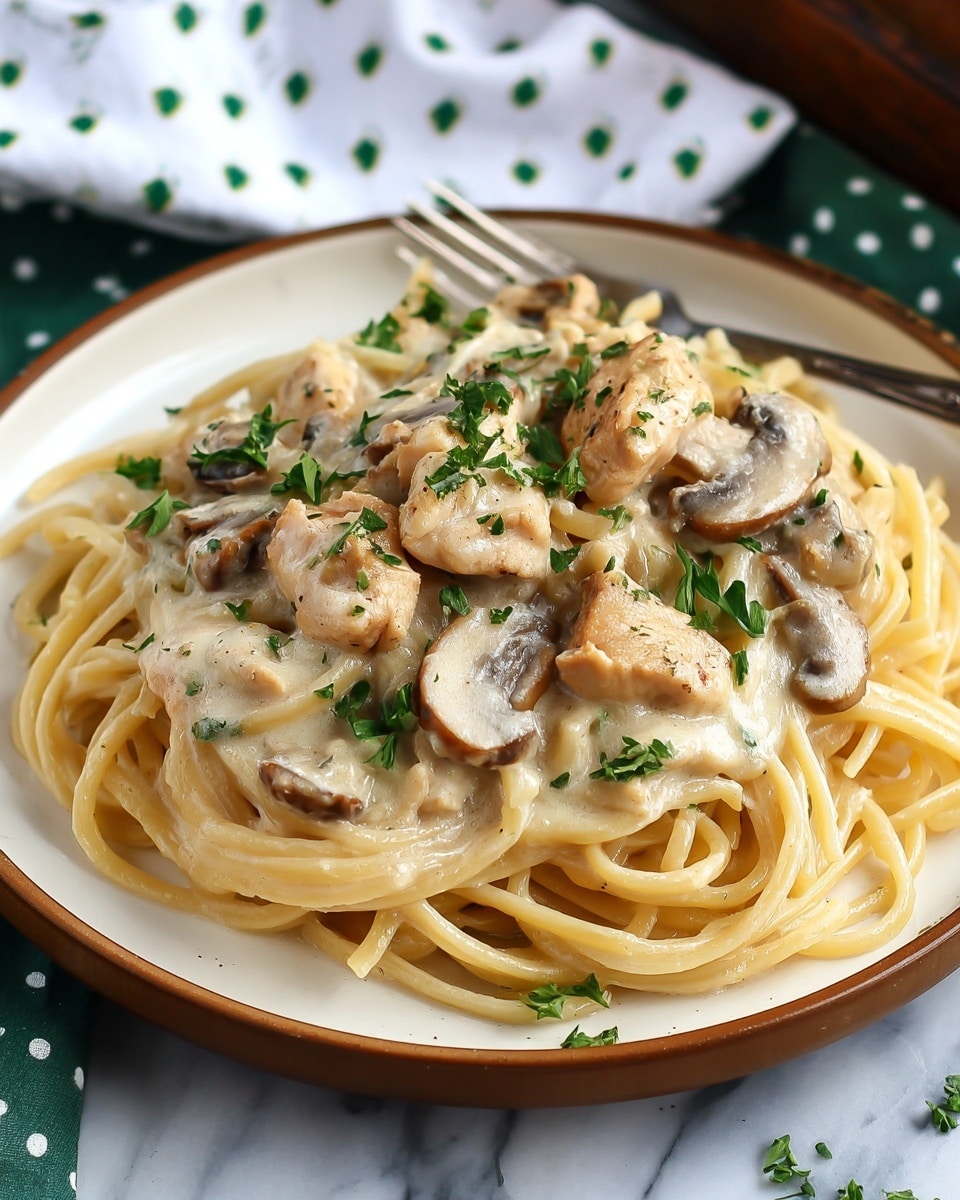 A dish of creamy chicken alfredo pasta is served on a white plate with a brown rim, placed on a white marbled surface. The bottom layer shows a bed of thick spaghetti noodles coated in a smooth, pale cream sauce. Scattered on top are pieces of browned, tender chicken chunks and sautéed mushroom slices, both with a slightly golden sear. Fresh green parsley is sprinkled over the dish, adding a touch of color contrast. In the background, a silver fork rests on the edge of the plate, and a white cloth with green dots is slightly visible. Photo taken with an iphone --ar 4:5 --v 7
