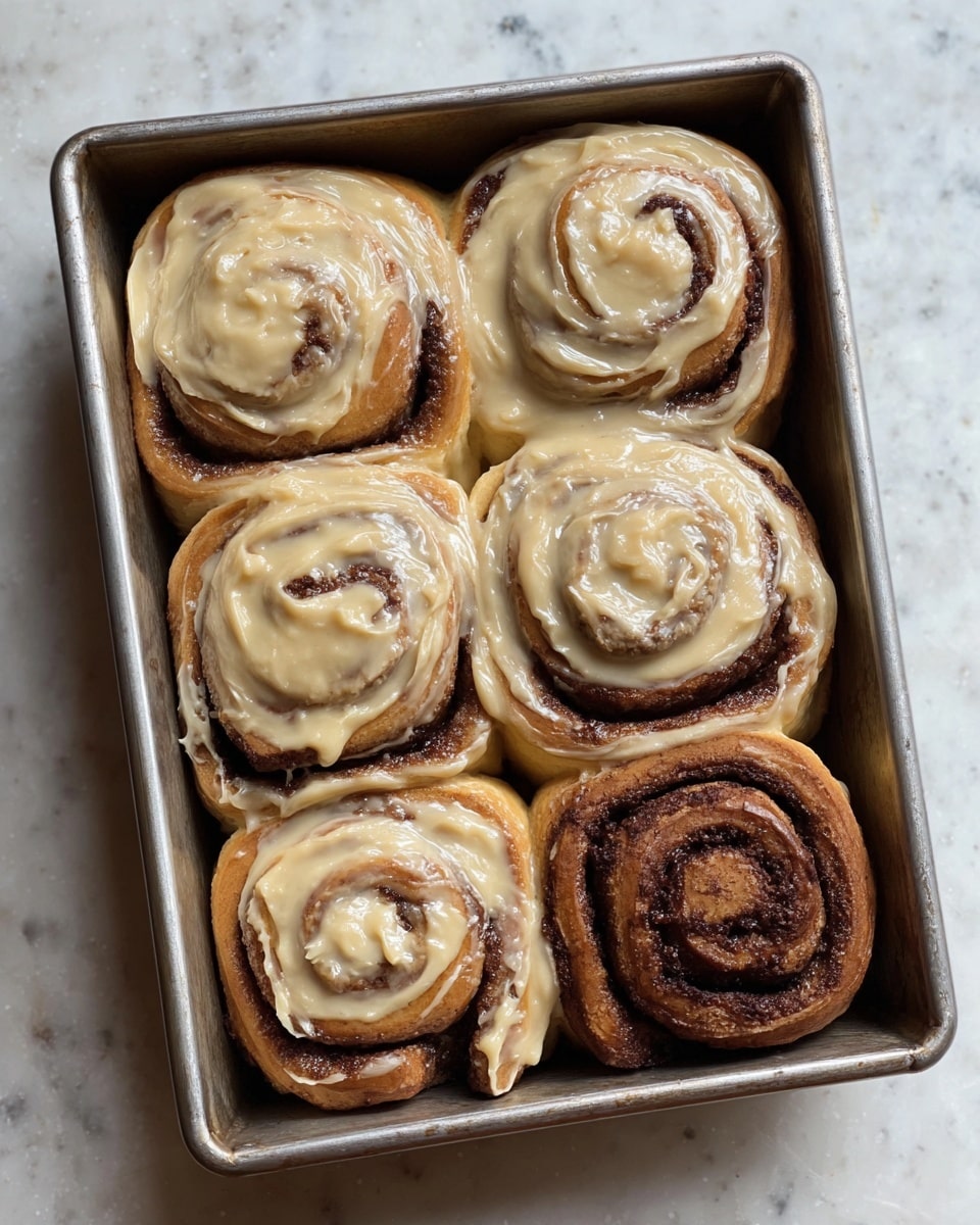 A metal baking tray holds six cinnamon rolls arranged closely in two rows of three. Five cinnamon rolls are thickly topped with a creamy, light caramel-colored frosting that covers almost the entire surface, with swirls of dark cinnamon visible underneath. The sixth cinnamon roll, located in the middle of the right column, is without frosting, showing a rich dark brown spiral pattern with a textured, soft dough look and some glistening sugar spots. The tray sits on a surface with a white marbled texture. photo taken with an iphone --ar 4:5 --v 7