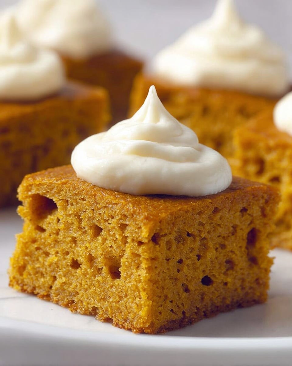 A close-up image of a single square piece of moist, spongy pumpkin cake with a golden brown crumb texture and small air holes visible throughout. The cake has a dollop of smooth, creamy white frosting piped on top in a swirl shape with a pointed peak. In the blurred background, there are other pieces of the same cake. The cake is set on a white plate placed on a white marbled surface. photo taken with an iphone --ar 4:5 --v 7