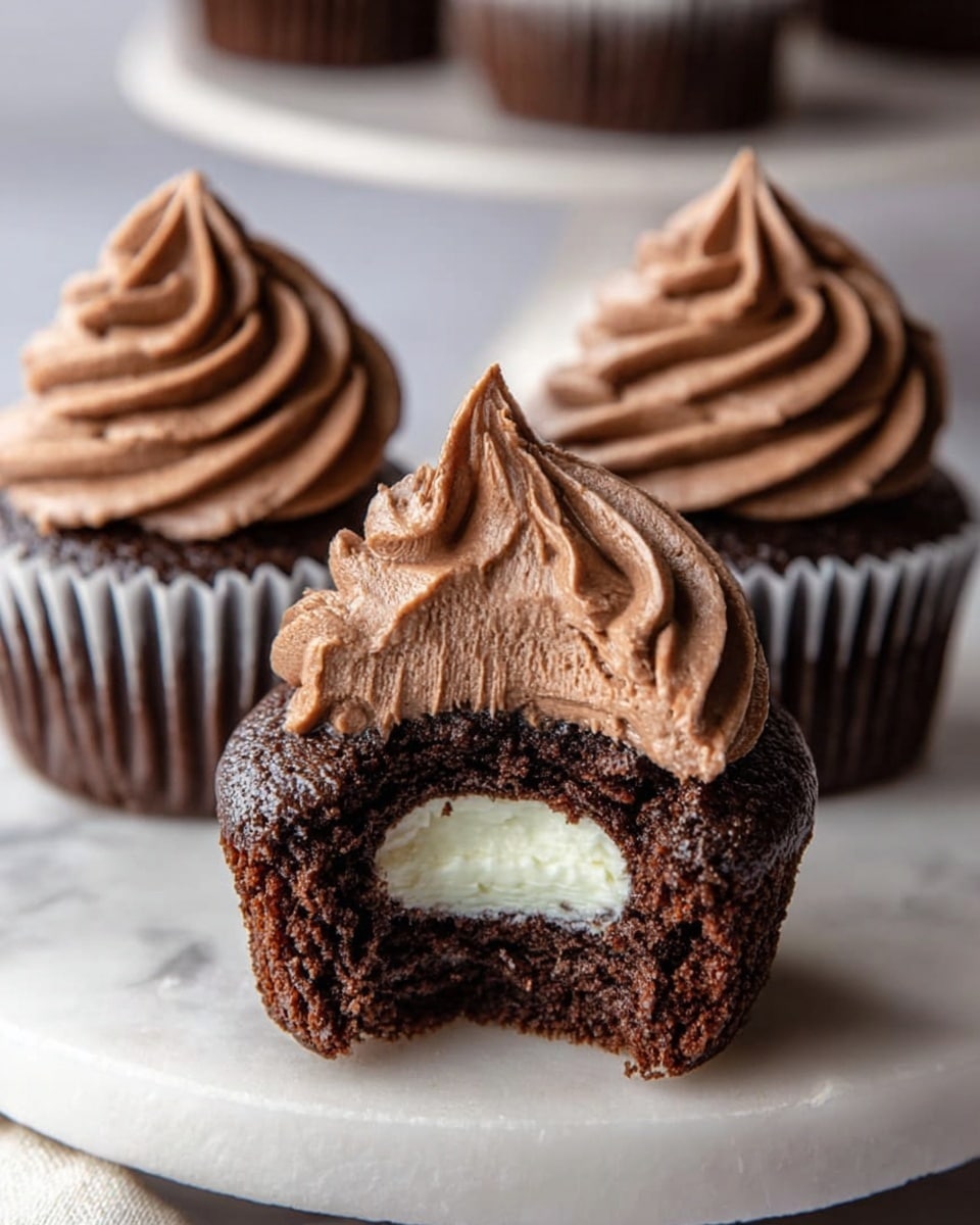 Three chocolate cupcakes are placed on a white round plate atop a white marbled surface. Each cupcake has a dark brown chocolate base with a white paper liner. The front cupcake has a bite taken out, revealing two layers inside: a middle layer of smooth chocolate cream and a center of white cream. On top of each cupcake is a swirl of rich, light brown chocolate frosting with soft peaks, the front cupcake’s swirl slightly indented to show the white cream inside. The background is softly blurred, focusing on the cupcakes. photo taken with an iphone --ar 4:5 --v 7