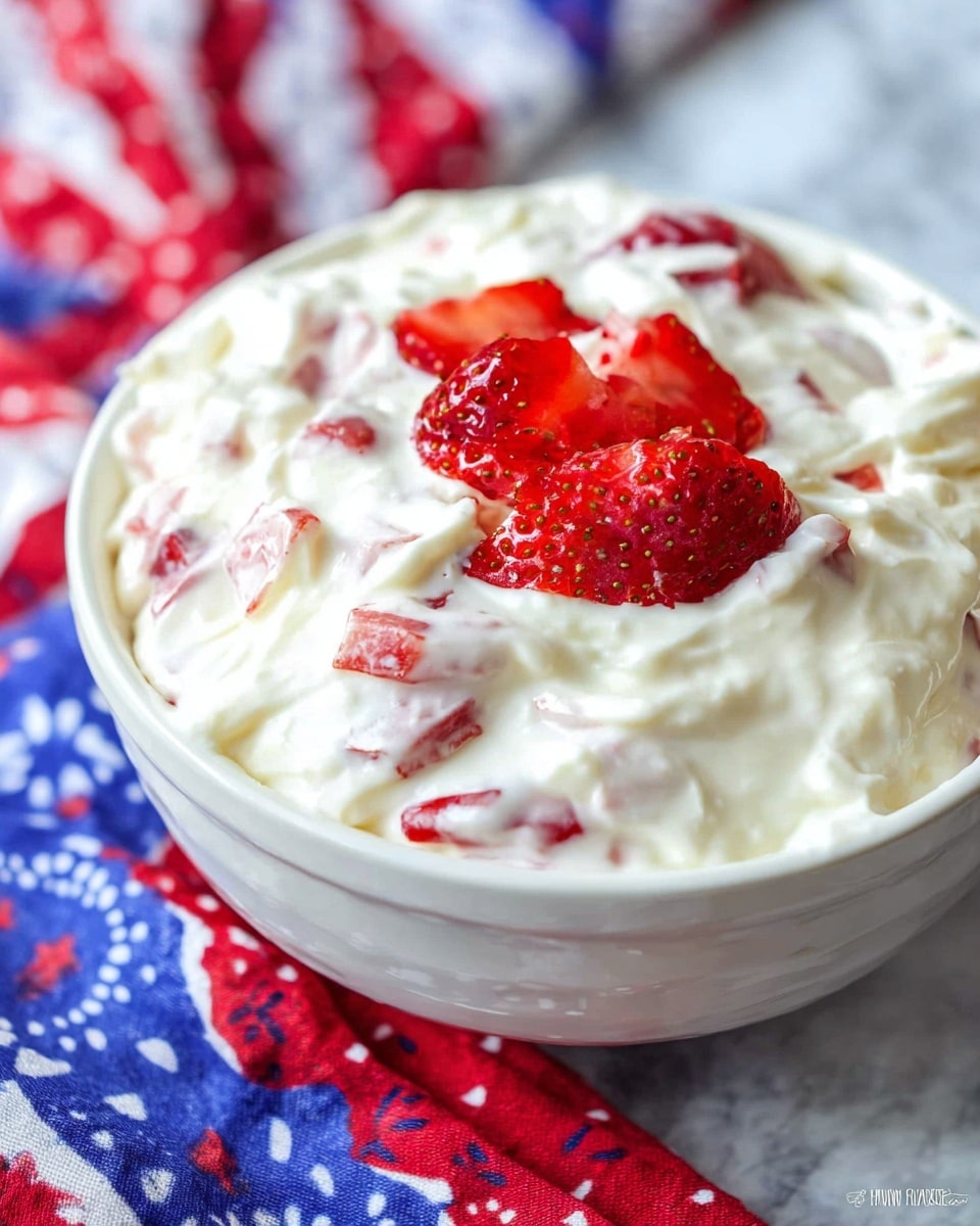 The image shows a close-up of a white bowl filled with a creamy dessert that has a soft, smooth texture. The dessert has visible layers of white cream mixed with small pieces of red strawberries scattered throughout the top and inside. The strawberries add bright red color and a juicy texture contrast to the thick, white cream. The bowl is placed on a surface covered with a white marbled texture, and parts of colorful, patterned cloths in red, blue, and white are seen near the bowl. Photo taken with an iphone --ar 4:5 --v 7