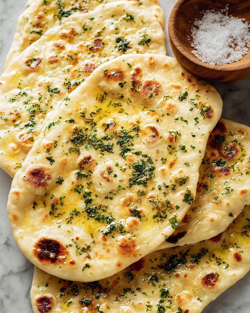 Several pieces of flatbread are stacked on a white marbled surface, each piece showing a soft, slightly puffy texture with golden-brown spots from cooking. The flatbreads have a light yellow layer of melted butter on their surface, sprinkled generously with finely chopped green herbs. A wooden bowl filled with coarse salt sits near the flatbreads, adding a rustic touch. The flatbreads are arranged overlapping each other, showing their irregular oval shapes and soft edges. photo taken with an iphone --ar 4:5 --v 7