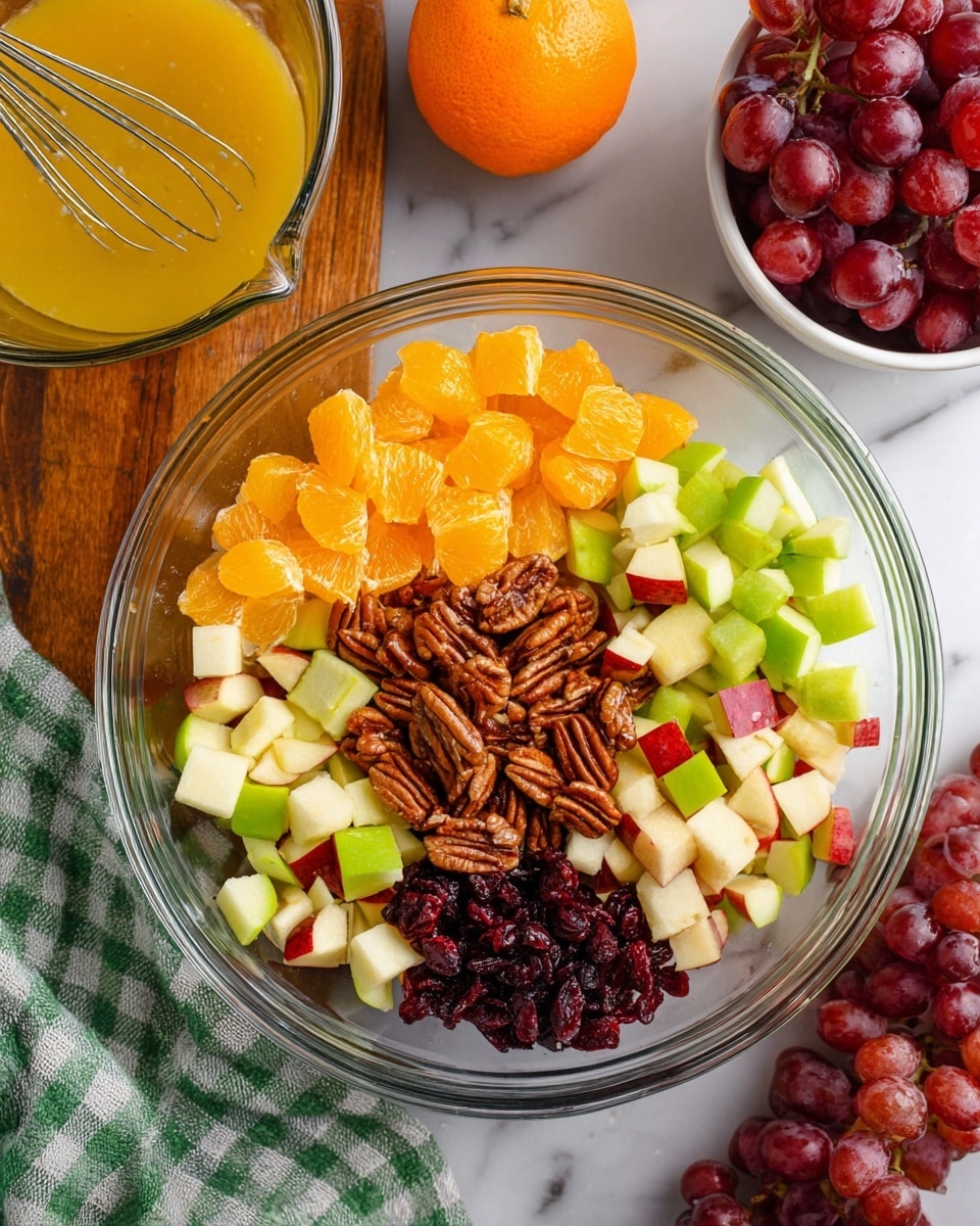 A clear glass bowl sits on a white marbled texture, filled with six sections of fresh fruit and nuts arranged separately: bright orange chunks of orange in the top left, pale green apple pieces on the right, white and red apple pieces on the left, red grapes at the bottom, a cluster of pecans in the center overlapping the grapes, and dark dried cranberries just above the pecans. To the top left of the bowl, a clear glass cup holds a yellow-orange dressing with a metal whisk inside. A whole orange fruit is placed near the top center. To the top right, a white bowl holds more red grapes. A green and white checkered cloth is draped near the bottom left of the image. photo taken with an iphone --ar 4:5 --v 7