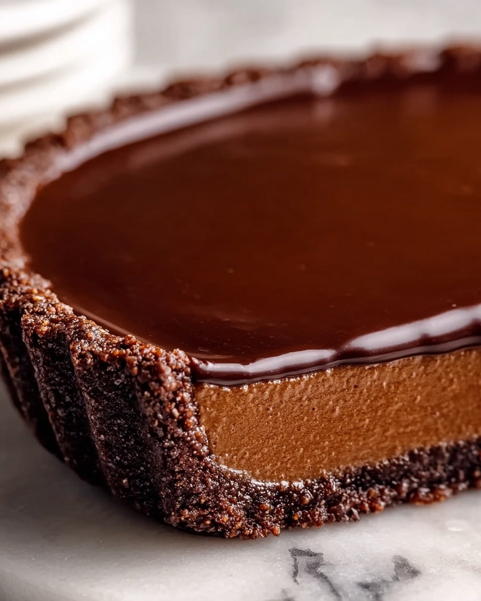 The image shows a chocolate tart with three distinct layers on a white marbled plate. The bottom layer is a dark brown crumbly crust, thick and firm, making the base and edges. The middle layer is a smooth, creamy milk chocolate filling with a lighter brown color and a velvety texture. The top layer is a glossy, dark chocolate ganache, which is shiny and reflective, covering the entire surface evenly. A piece of the tart is missing, revealing the clean cut of all three layers. The plate is on a white marbled surface with some small chocolate pieces nearby. photo taken with an iphone --ar 4:5 --v 7