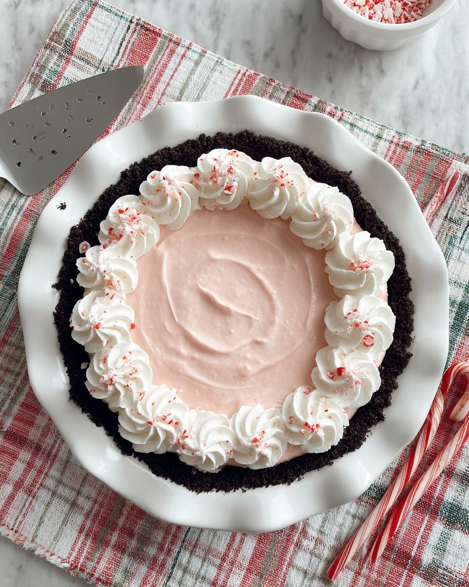 A dessert pie is shown on a white scalloped-edge plate set on a white marbled texture with a red, green, and white checkered cloth underneath. The pie has a crumbly, dark chocolate crust forming the bottom and sides. The middle layer is smooth, pale pink filling, spread evenly with light swirls visible on the surface. On top, a ring of white whipped cream rosettes is piped around the edge of the pink filling, each rosette sprinkled with small red bits that resemble crushed candy canes. A silver slotted spatula is placed near the top left, two candy canes lie crossed on the bottom left, and a white bowl sits slightly out of focus on the top right. A red and white striped cloth is partially visible on the right side. photo taken with an iphone --ar 4:5 --v 7