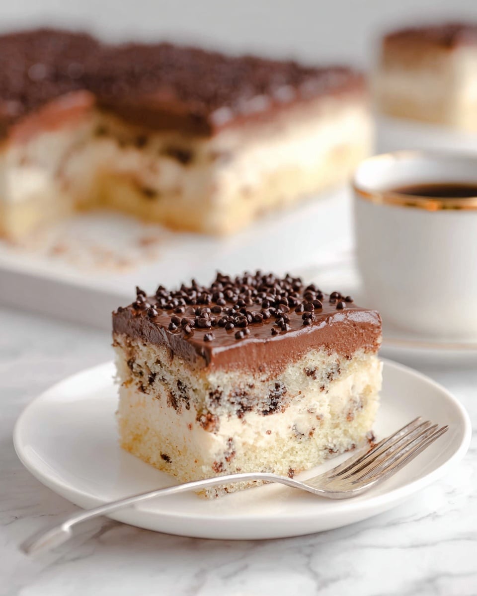 The image shows a square piece of cake on a white plate with a fork beside it. The cake has three visible layers: the bottom and middle layers are light beige cake with small dark chocolate chips inside, and the top layer is smooth, thick dark chocolate frosting sprinkled with mini chocolate chips. In the background, a larger rectangular cake with the same layers is slightly out of focus, resting on a white marbled surface. To the side, there is a white cup with a gold rim, partially visible. photo taken with an iphone --ar 4:5 --v 7