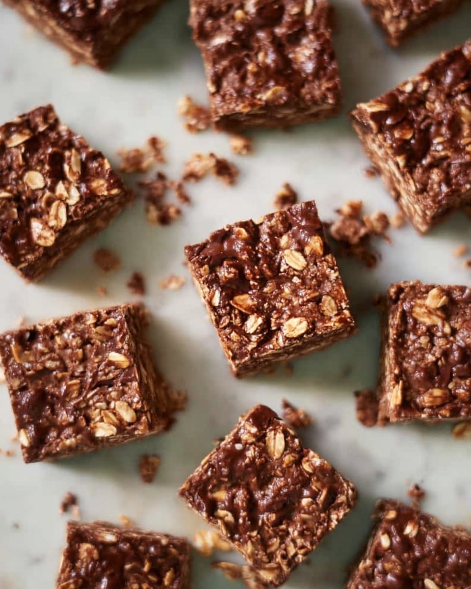 The image shows multiple small, square-shaped chocolate oat bars arranged closely together on a white marbled surface. Each bar has a rough texture with visible oats embedded in a dark brown chocolate base, giving a mix of shiny and matte spots. The bars are unevenly spaced, some with crumbs scattered around them, adding to the homemade look. The squares appear thick and dense, highlighting the oats on top and pockets of chocolate throughout. photo taken with an iphone --ar 4:5 --v 7