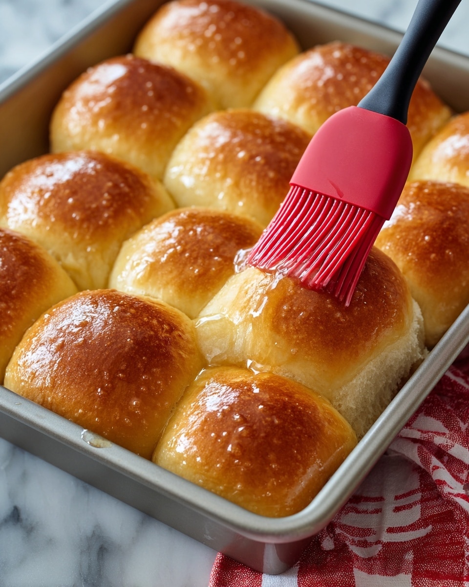 The image shows a close-up of a square metal baking pan filled with 16 soft, golden brown dinner rolls arranged in a 4 by 4 grid. Each roll has a smooth, slightly shiny surface with a warm beige color that darkens to a light golden brown at the top. A red silicone brush with a black handle is applying a glossy glaze to the top of one roll in the center-right area, making it look moist and shiny. The pan sits on a white marbled surface with a glimpse of a red and white cloth nearby. photo taken with an iphone --ar 4:5 --v 7