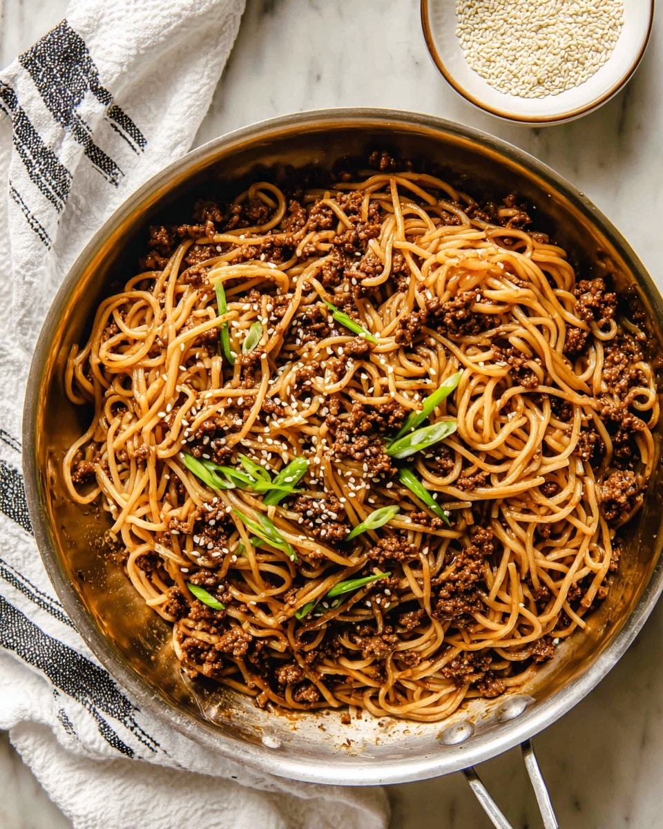 This image shows a silver pan filled with a single-layer of noodles mixed with cooked ground meat that is dark brown and evenly distributed. The noodles are light brown and glossy from the sauce, slightly tangled. Scattered on top are small white sesame seeds and thin green rings of chopped scallions adding fresh color. To the upper right of the pan is a white bowl filled with white sesame seeds. The pan sits on a white marbled surface with a white cloth with black stripes underneath its handle. Photo taken with an iphone --ar 4:5 --v 7