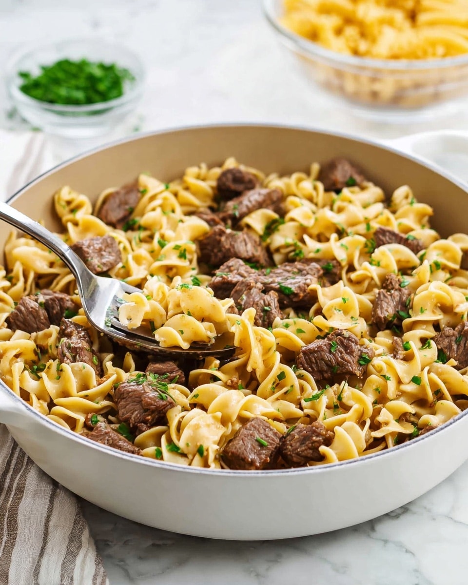 A white pan filled with cooked egg noodles mixed with small, browned beef chunks and sprinkled with chopped green herbs evenly over the top. The noodles are soft and light yellow, swirling around the tender beef pieces which have a rich brown color. A silver fork rests inside the pan, lifting some noodles and beef. The background is a white marbled surface with some fresh green herbs and a blurred bowl of dry pasta in the distance, creating a fresh, clean setting. photo taken with an iphone --ar 4:5 --v 7