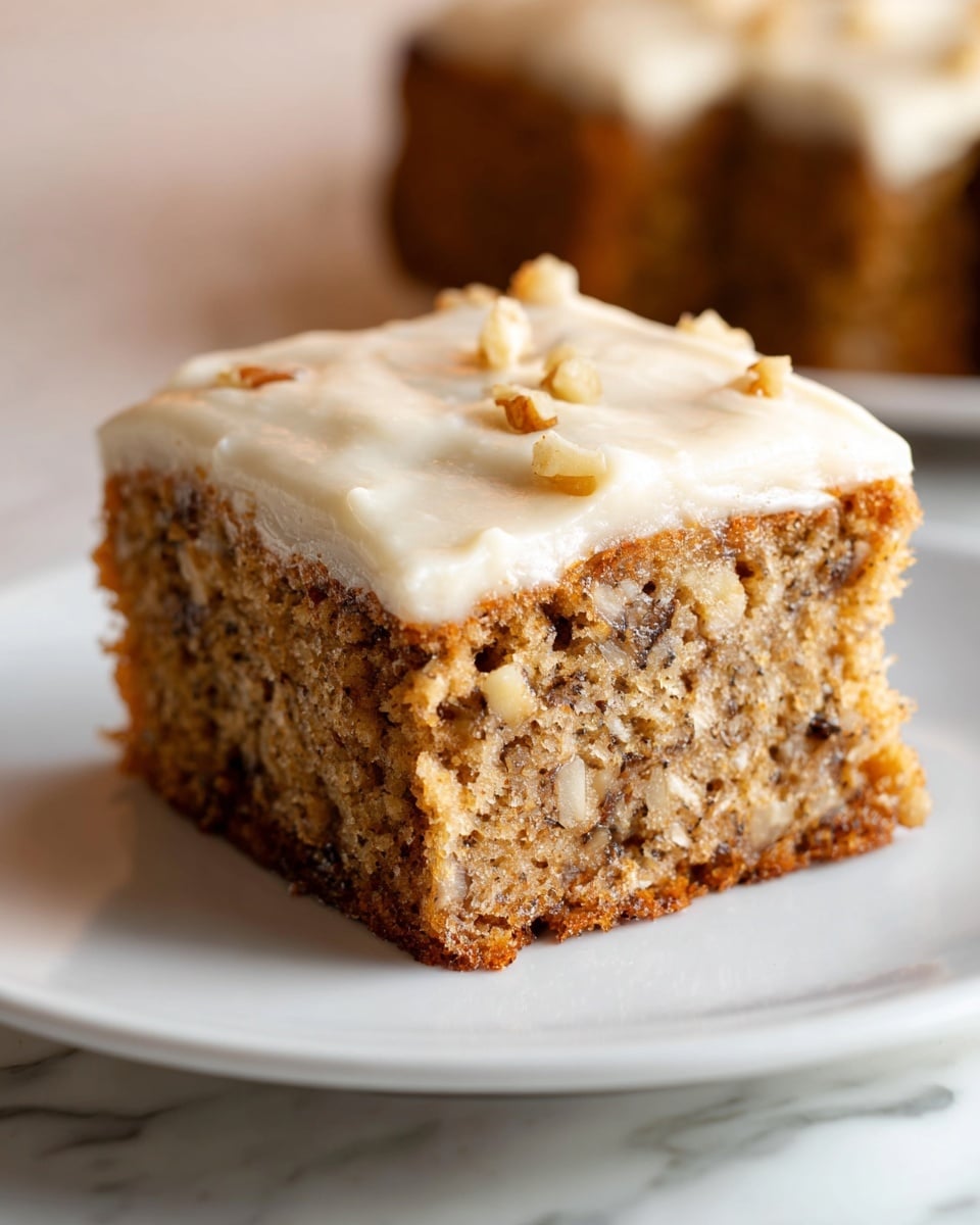 A single square piece of nut cake with moisture and crumb texture is placed on a white plate, showing a thick cream-colored frosting covering the entire top layer smoothly with a few small nut pieces embedded. The cake layer is light brown and dense, filled with small nut chunks and dark spots inside. The plate sits on a surface with a white marbled texture, and the background is soft and blurred, keeping focus on the cake. Photo taken with an iphone --ar 4:5 --v 7