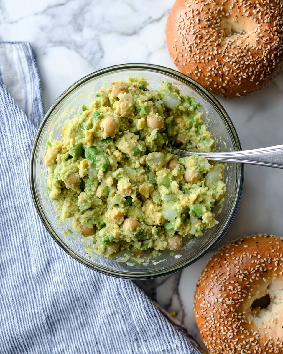 A clear glass bowl filled with a chunky mix of light green avocado pieces and pale yellow chickpeas, showing a slightly mashed texture with small bits throughout, and a silver spoon resting inside. Next to the bowl are two sesame seed bagels with a shiny golden brown crust and visible white sesame seeds, placed on a white marbled surface partly covered by a blue and white striped cloth. Photo taken with an iphone --ar 4:5 --v 7