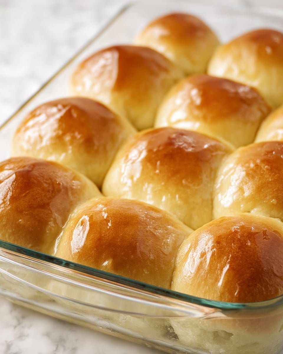 A glass baking dish filled with nine soft, round bread rolls that are golden brown on top with a shiny, slightly glazed texture. The rolls are touching each other and appear fluffy with a smooth crust. The dish is placed on a white marbled textured surface. The lighting highlights the warm color and soft texture of the bread. photo taken with an iphone --ar 4:5 --v 7