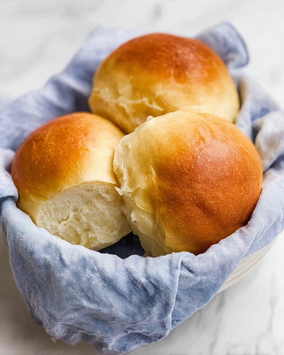 Three soft, golden brown bread rolls sit close together inside a white bowl lined with a light blue cloth. The bread rolls have a smooth, shiny crust on top with a soft, fluffy texture visible on the sides and inside where one roll is torn slightly. The cloth is gently folded, adding a cozy feel to the setting. The bowl is on a white marbled surface. photo taken with an iphone --ar 4:5 --v 7