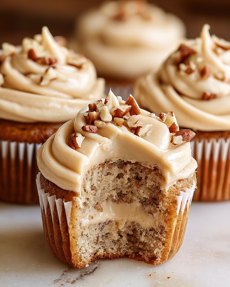 A close-up view of a moist cupcake with a bite taken out, revealing a soft, light brown inner layer with a creamy beige filling in the center. The top is covered with a thick swirl of smooth, pale cream frosting, decorated with chopped pecan nuts scattered on top. The cupcake is in a white paper liner, sitting on a white marbled surface, with a few more cupcakes and pecan pieces blurred in the background. photo taken with an iphone --ar 4:5 --v 7