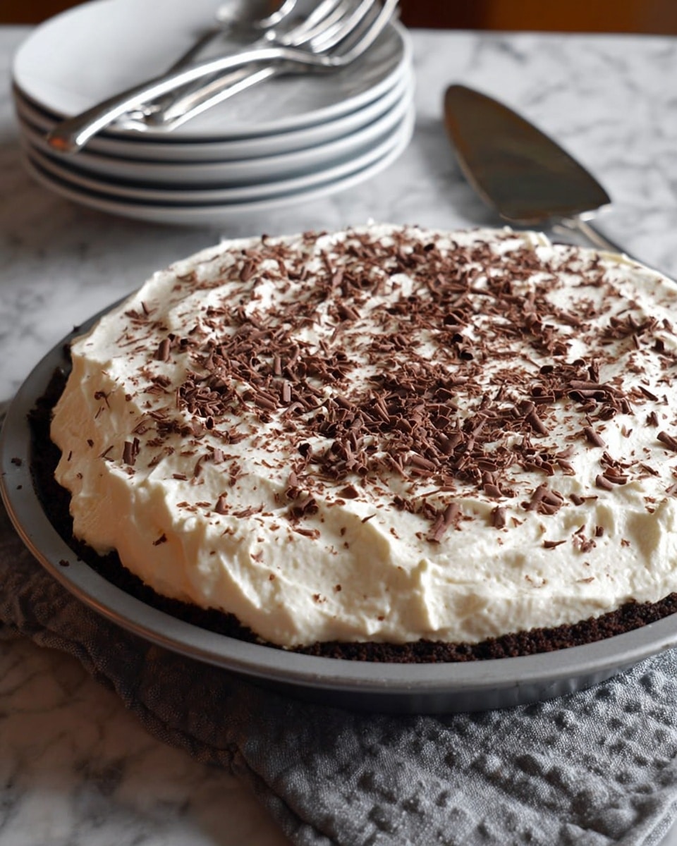 A round pie sits on a white marbled surface in a gray metal pie dish. The pie has a thick, fluffy white whipped cream layer spread evenly on top, sprinkled generously with small dark brown chocolate shavings. The edges of the whipped cream layer are slightly uneven and soft, creating a light texture contrast with the pie's base, which is barely visible but dark brown, suggesting a chocolate or cookie crust beneath. In the background, there is a stack of white plates with silver forks resting on top, all placed on the same white marbled surface. Near the pie dish, a shiny silver pie server is partially visible. photo taken with an iphone --ar 4:5 --v 7