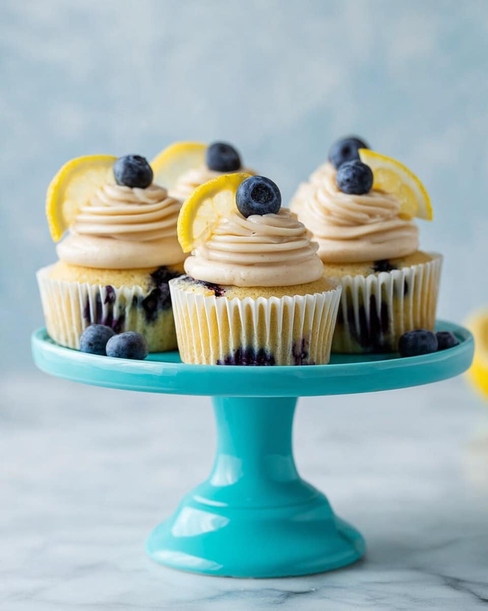Four cupcakes each have a base layer of yellow cake mixed with visible dark blueberries inside. On top, there is a light beige, thick swirled frosting made of three large round layers. Each cupcake is decorated with a single dark blue, round blueberry on top and a thin curved yellow lemon slice placed behind the blueberry. The cupcakes are held in white paper liners. They are placed on a bright turquoise cake stand with a wide scalloped edge and a smooth column base. The background is a soft white marbled texture. Photo taken with an iphone --ar 4:5 --v 7