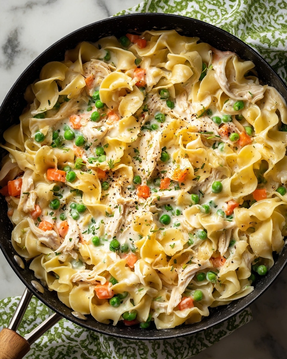 A close-up view of a creamy pasta dish in a black pan, showing a mix of three layers: wide egg noodles with soft, curly edges in a pale yellow color, small bright green peas, and diced soft orange carrots scattered evenly throughout. Shredded pieces of light beige chicken are mixed in, all coated with a smooth, creamy white sauce that has a slightly glossy texture and sprinkled black pepper dots. The pan is placed on a white marbled surface with a green and white patterned cloth partially visible. Photo taken with an iphone --ar 4:5 --v 7