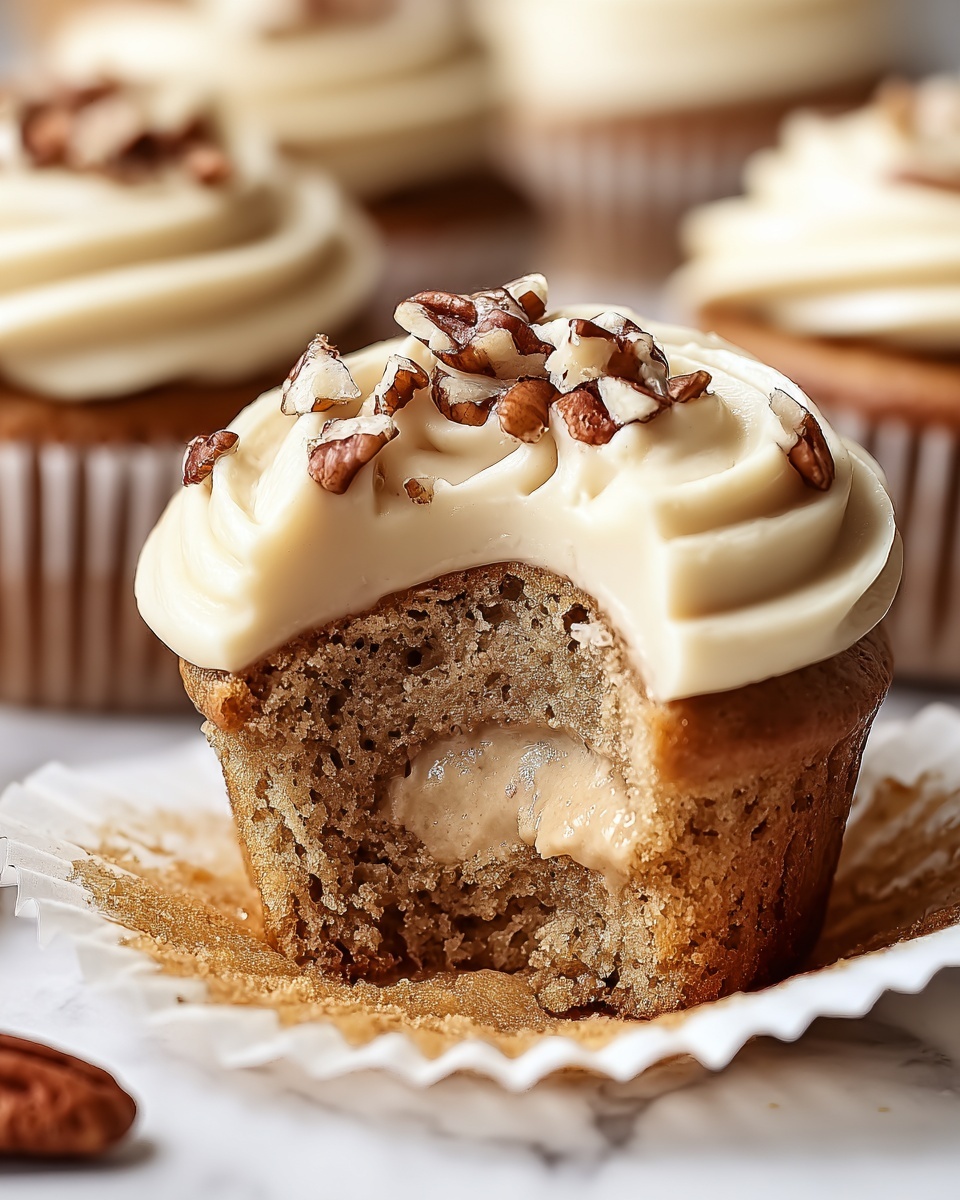 Three cupcakes sit on a white marbled textured surface, each topped with smooth, creamy light beige frosting swirled neatly in two thick layers. The cupcake in the front has a bite taken out, revealing a soft, moist, speckled light brown cake with a middle layer of creamy beige filling. The frosting is sprinkled with pieces of chopped pecans in golden brown shades. The white cupcake liners hug the base of each cupcake. The blurred background adds depth but keeps focus on the detailed texture and layers of the cupcakes. photo taken with an iphone --ar 4:5 --v 7