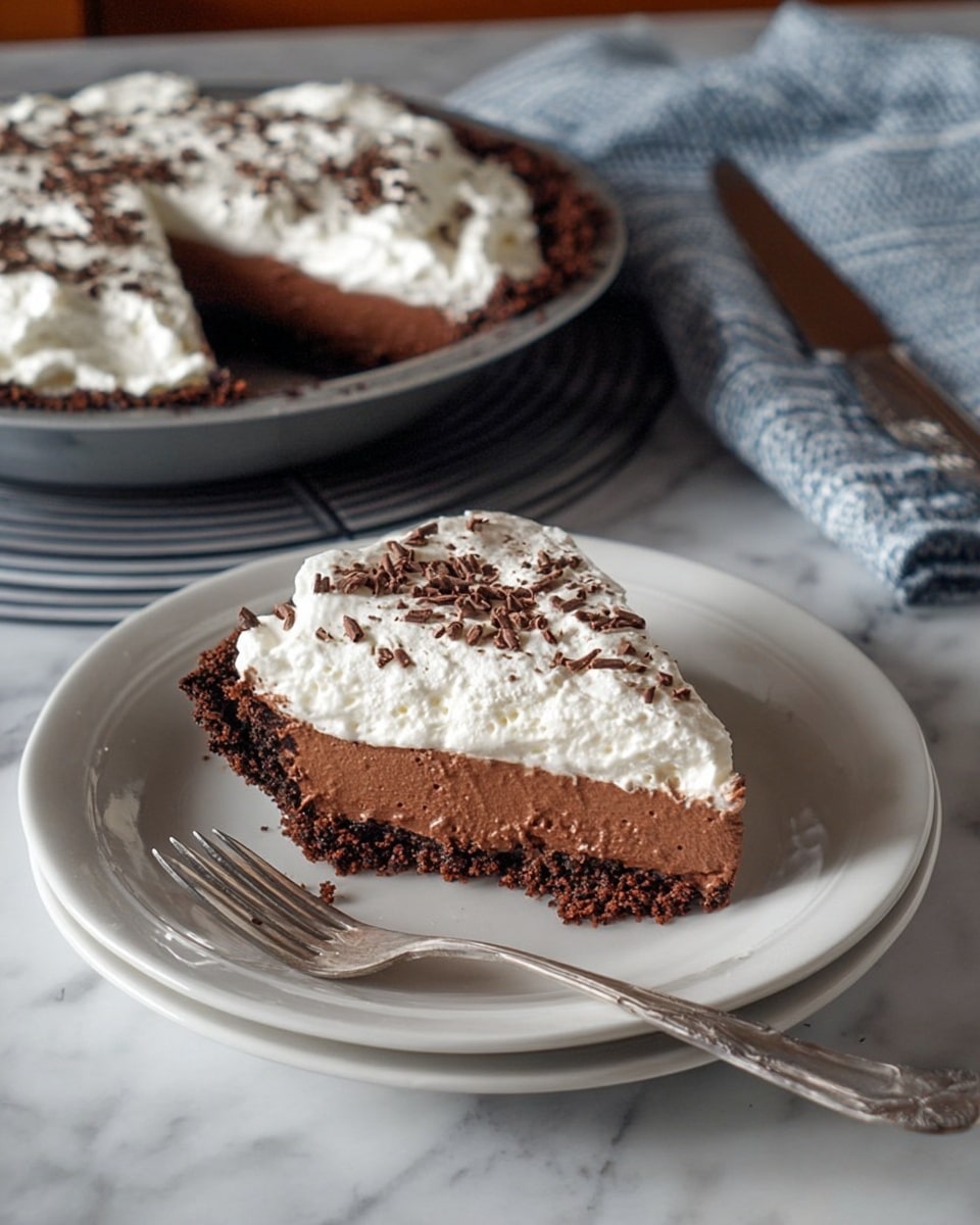 The image shows a slice of chocolate cream pie on a white plate, placed on a white marbled surface. The pie has three visible layers: the bottom is a dark brown chocolate crust with a crumbly texture, the middle is a thick, smooth chocolate cream layer with a rich dark color, and the top layer is a fluffy white whipped cream sprinkled with small dark chocolate shavings. In the background, the rest of the pie sits in a grey pan on a dark round charger plate, with one slice missing. A silver fork rests to the left of the slice on the plate, and a knife and a blue and white striped cloth are seen in the upper right part of the image. photo taken with an iphone --ar 4:5 --v 7
