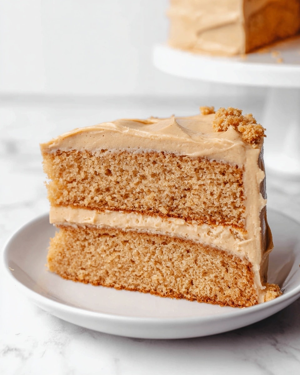 A slice of cake with two thick layers of light brown, soft crumb shows a smooth, light tan frosting spread evenly on top and between the layers. There are small pieces of crumb sprinkled on the top edge. The slice sits on a simple white plate, set against a clean white marbled surface in the background. Photo taken with an iphone --ar 4:5 --v 7