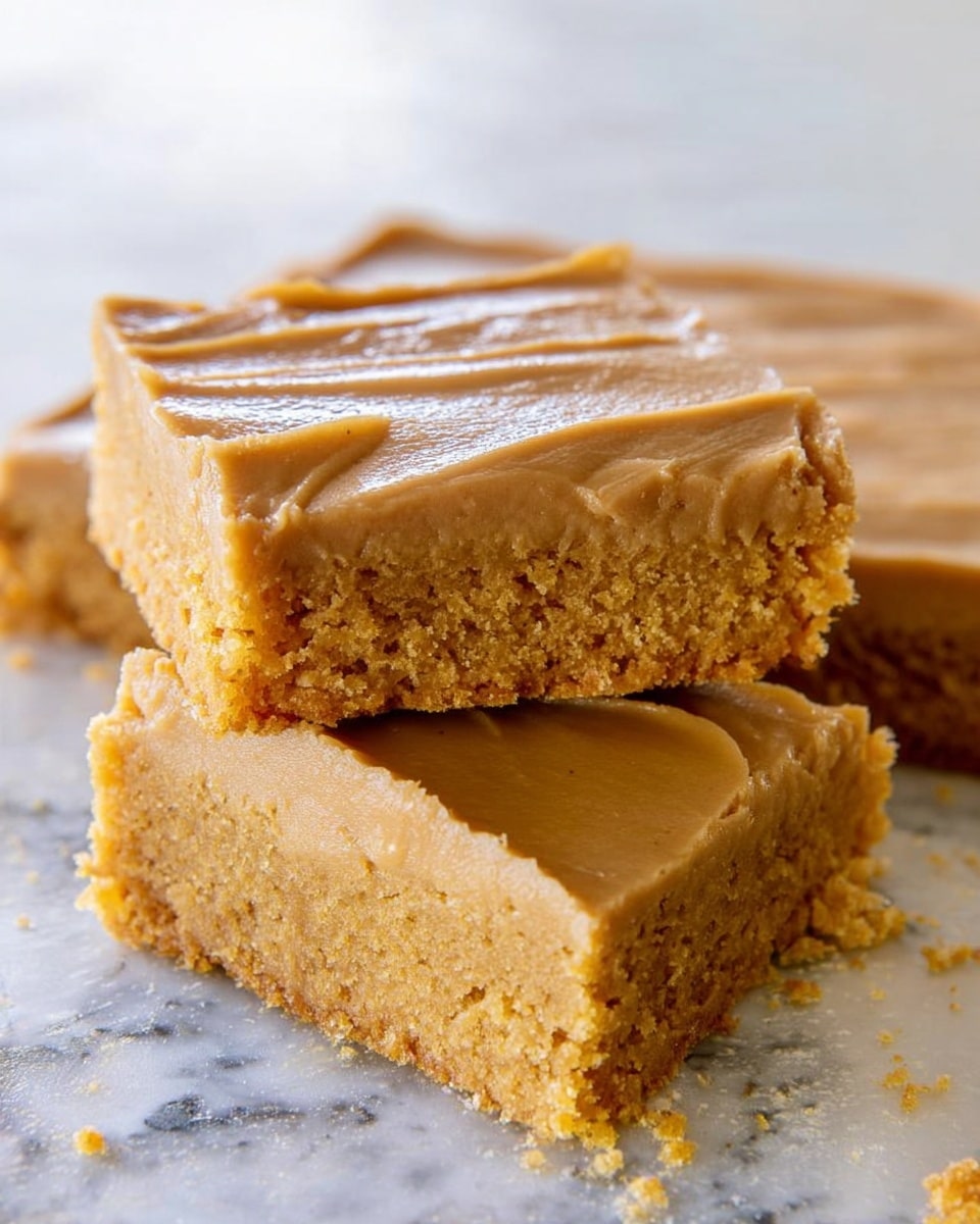 The image shows two square pieces of a dessert with two distinct layers. The bottom layer is a crumbly, golden-brown cake with a moist texture. The top layer is a smooth, light brown frosting with a glossy finish and a soft spread pattern. The upper piece is stacked slightly off-center on the lower piece, revealing the thick cake base beneath. The background is a white marbled texture with some crumbs scattered around the dessert. Photo taken with an iphone --ar 4:5 --v 7