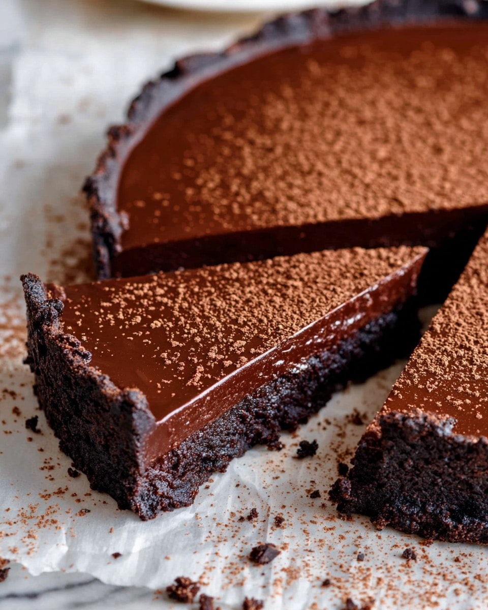 A rich chocolate tart sliced into pieces, showing two clear layers: the bottom layer is dark, dense, and crumbly with a rough texture, while the top layer is smooth, glossy chocolate ganache with a shiny finish; the surface of the ganache is dusted with cocoa powder that adds a light brown speckled effect; the tart sits on white parchment paper which is placed on a white marbled texture, with a few crumbs scattered around the tart edges, creating a slightly messy yet inviting look. photo taken with an iphone --ar 4:5 --v 7