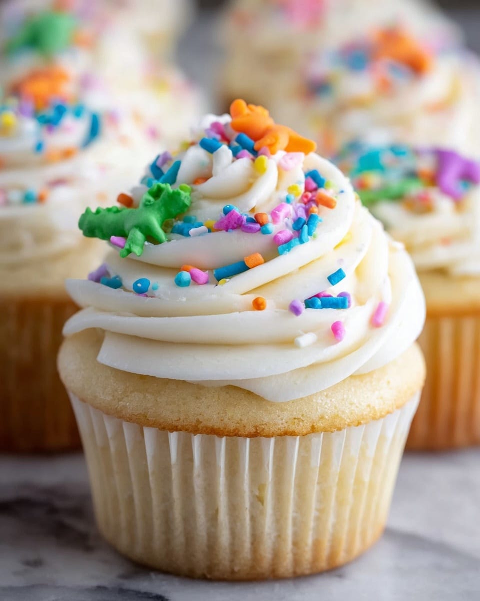 A close-up of a white cupcake with a bite taken out of it, showing a soft, fluffy white cake base filled with colorful sprinkles inside. The cupcake is topped with a thick layer of smooth white frosting, decorated with bright, multicolored sprinkles and small shaped candy pieces, including a green cactus and an orange animal figure on top. The cupcake liner is white, and the background is a white marbled texture. photo taken with an iphone --ar 4:5 --v 7