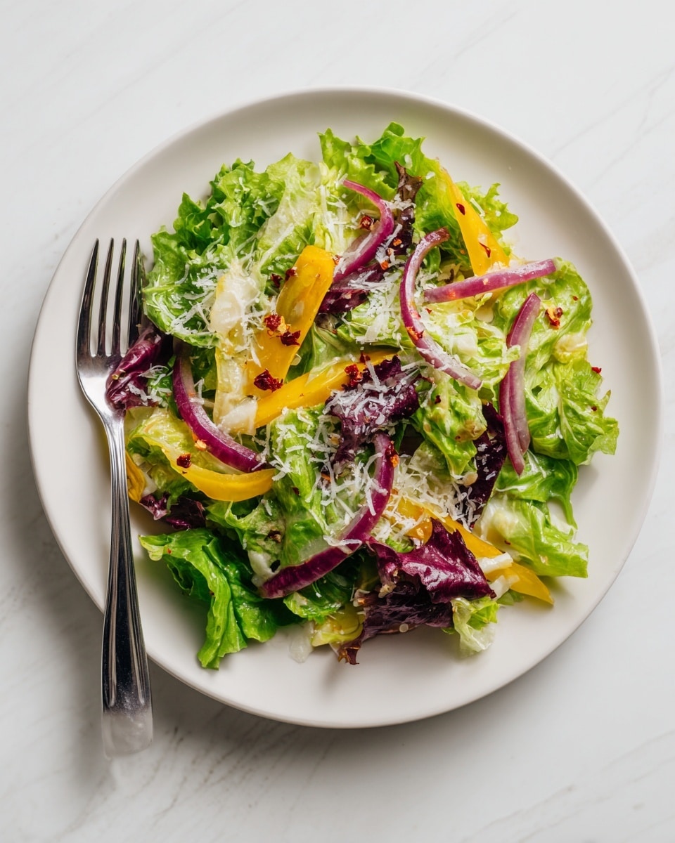 A fresh salad is shown on a white plate placed on a white marbled surface. The salad is made of roughly chopped green and yellow leafy lettuce as the base layer with thin, curved slices of purple onion scattered throughout. The top has a light dusting of grated cheese and small red chili flakes. A silver fork is resting on the left side of the plate. Photo taken with an iphone --ar 4:5 --v 7
