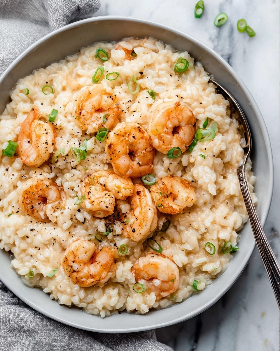 A bowl filled with creamy risotto rice, cooked with small tender shrimp scattered evenly throughout. The rice is soft and pale beige, with a slightly glossy and smooth texture, mixed with bits of green onion slices sprinkled on the top. The plump shrimp are light pink with a slight char and a firm texture, some resting on top and others mixed inside the risotto. A few black pepper specks are sprinkled over everything, adding contrast. A silver spoon is placed inside the bowl, ready to scoop the dish. The bowl is white, sitting on a white marbled surface with a folded light gray cloth nearby. photo taken with an iphone --ar 4:5 --v 7