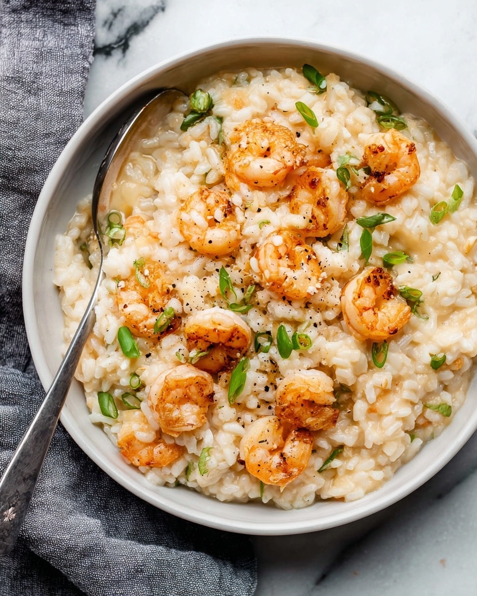 A close-up of a white bowl filled with creamy risotto rice that is light beige in color and has a soft, slightly glossy texture. On top and mixed throughout the rice are small, cooked shrimp with a pink-orange color and a slight char, giving them a grilled look. Scattered green pieces of thinly sliced scallions add a fresh bright contrast to the creamy risotto, while a light sprinkle of black pepper is visible on the surface. A silver spoon rests inside the bowl, partially submerged in the risotto, and the bowl sits on a white marbled surface with a folded grey cloth nearby. photo taken with an iphone --ar 4:5 --v 7