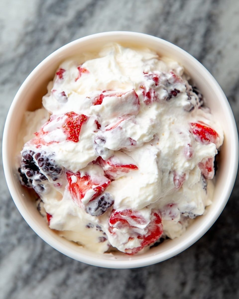 The image shows a white bowl filled with a creamy mixture that looks soft and fluffy. Inside the cream, there are visible pieces of red strawberries and dark purple blackberries evenly mixed throughout. The creamy layer is white and thick, surrounding the colorful fruit pieces, creating a contrast of red, purple, and white. The bowl sits on a white marbled surface, with the cream and fruit layers blending smoothly but distinct in color and texture. Photo taken with an iphone --ar 4:5 --v 7
