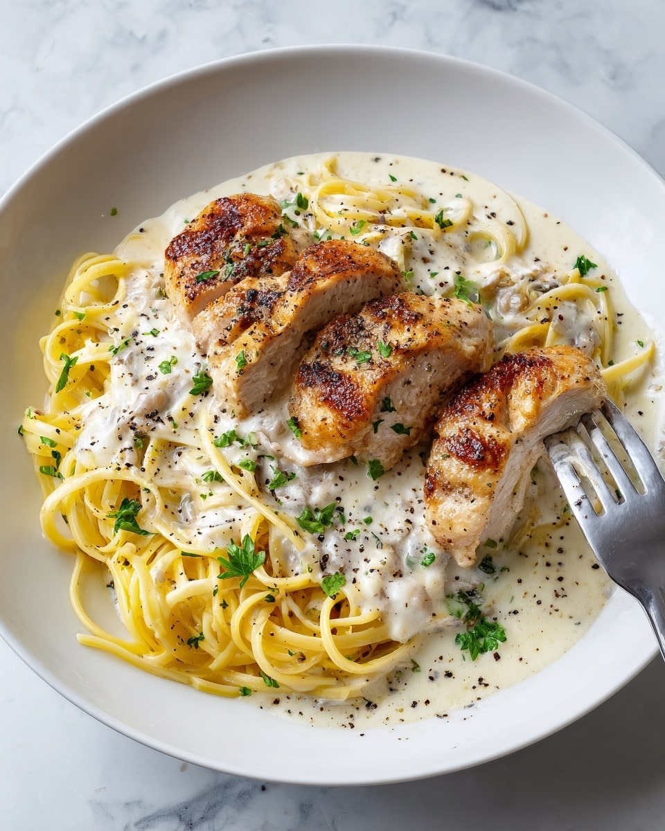 The dish is served in a white plate placed on a white marbled surface, showing a bed of yellow cooked spaghetti pasta as the bottom layer, with a creamy white sauce coating the noodles evenly. On top, there are two pieces of golden-brown, grilled chicken breasts with a lightly crispy, slightly charred texture, both covered partly in the creamy sauce. The dish is sprinkled generously with cracked black pepper and small chopped green parsley leaves, adding touches of black and bright green color. A silver fork is gently lifting a small portion of pasta towards the right side of the plate. photo taken with an iphone --ar 4:5 --v 7