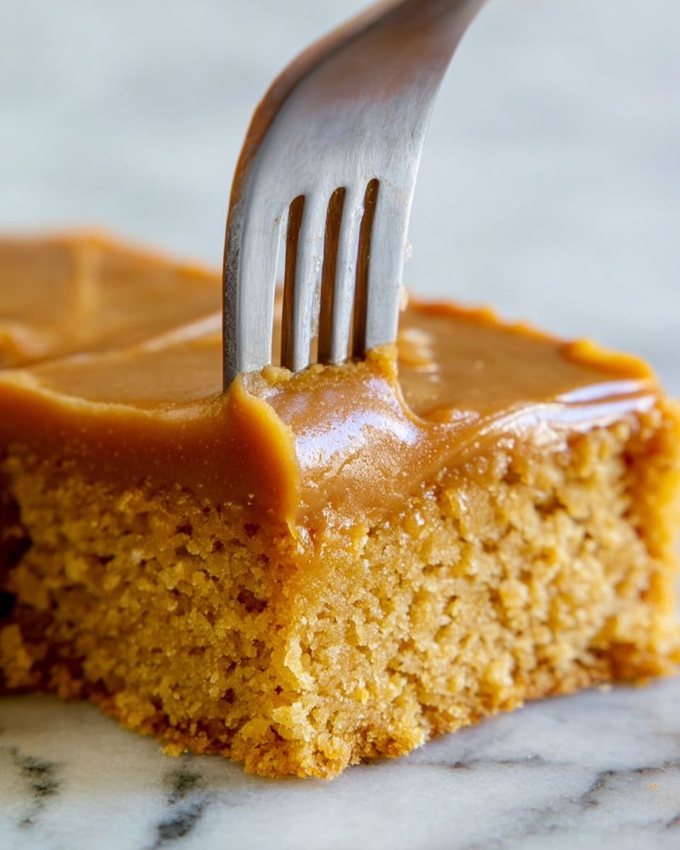 The image shows a close-up of a square piece of two-layer cake. The bottom layer is a crumbly, moist golden-brown cake with a textured surface. The top layer is a smooth, thick caramel-colored frosting, evenly spread and shiny. A metal fork with four tines is pressing down slightly into the frosting, creating a small indentation. The background has a white marbled texture. photo taken with an iphone --ar 4:5 --v 7