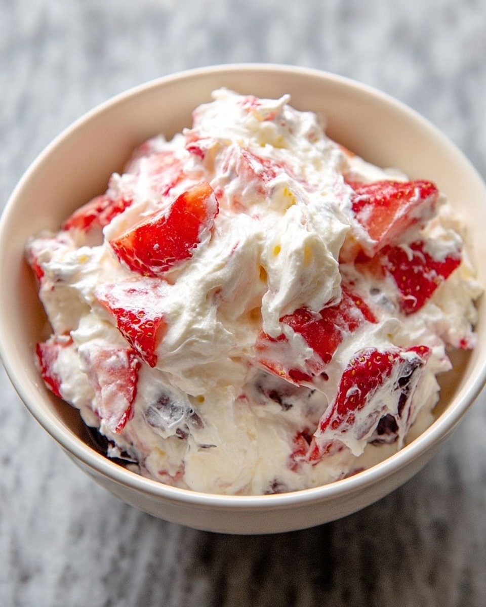 A close-up of a white bowl filled with a creamy dessert made of whipped cream mixed with red strawberries sliced showing their bright red color, along with some darker berries that appear to be blueberries, all blended into the thick, fluffy white cream in multiple layers. The mixture looks soft and airy with bits of fruit showing through the creamy texture, set on a white marbled surface. Photo taken with an iphone --ar 4:5 --v 7