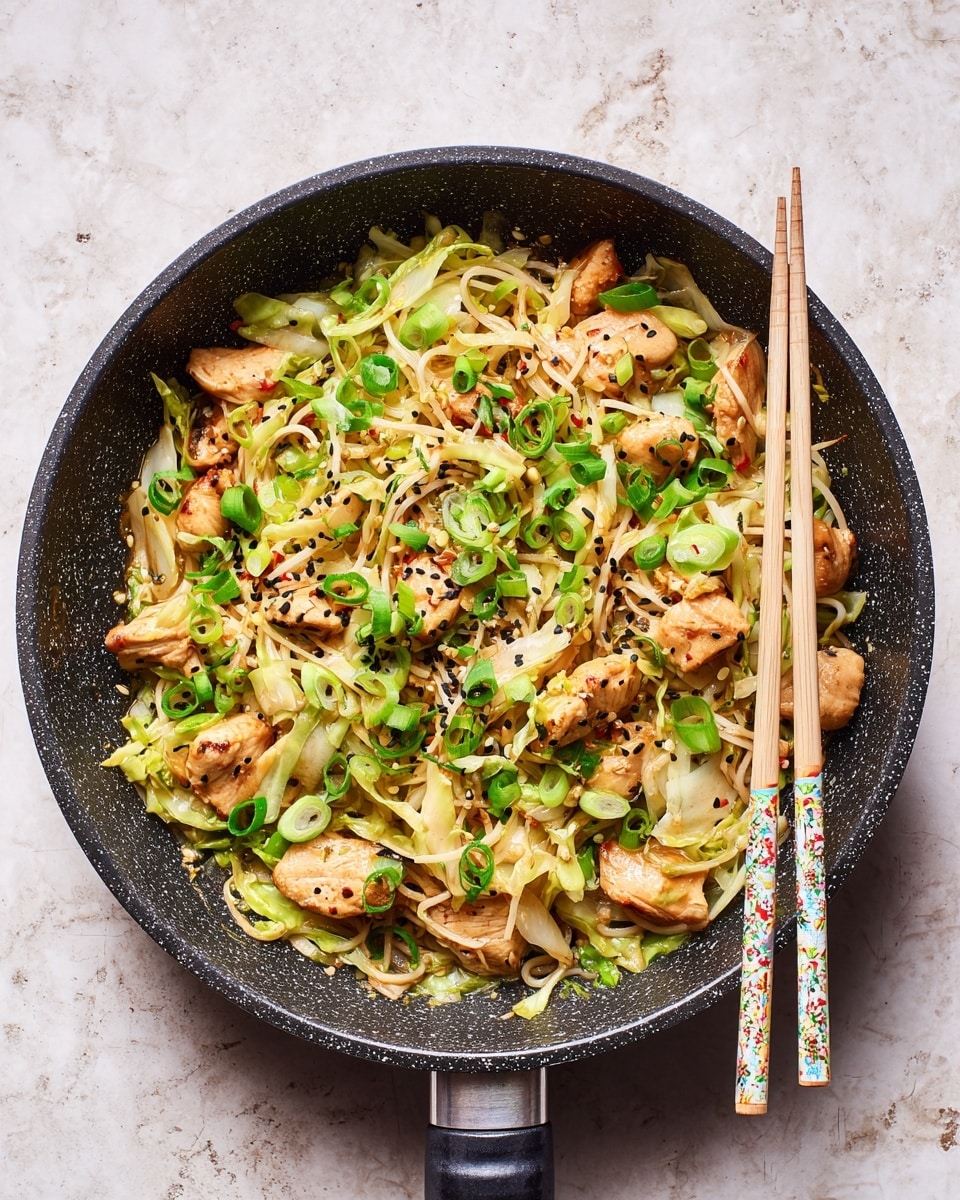A black speckled frying pan filled with a stir-fry dish consisting of light brown cooked chicken pieces mixed with pale green and white cabbage strips, topped with bright green chopped scallions and sprinkled with black and white sesame seeds and red chili flakes. The pan is placed on a white marbled textured surface, and a pair of light-colored chopsticks with a colorful pattern rests on top of the dish. photo taken with an iphone --ar 4:5 --v 7