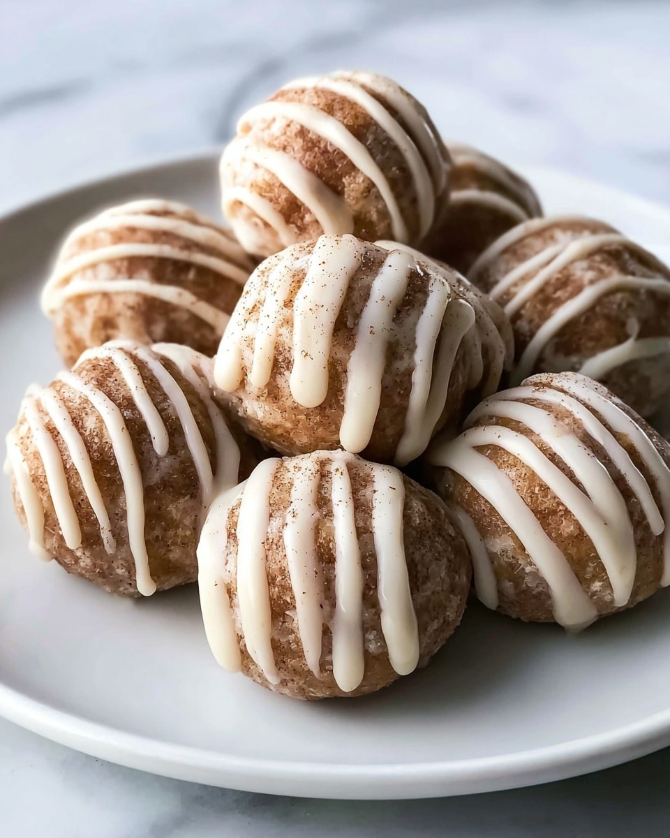 A close-up image shows a white plate filled with eight round, brown cinnamon balls. Each ball is covered in a light cream-colored glaze, with thicker cream lines drizzled over the top in a zigzag pattern. The brown balls have a slightly grainy texture with visible cinnamon specks. The scene is set on a smooth white marbled surface, giving a clean and simple look. photo taken with an iphone --ar 4:5 --v 7
