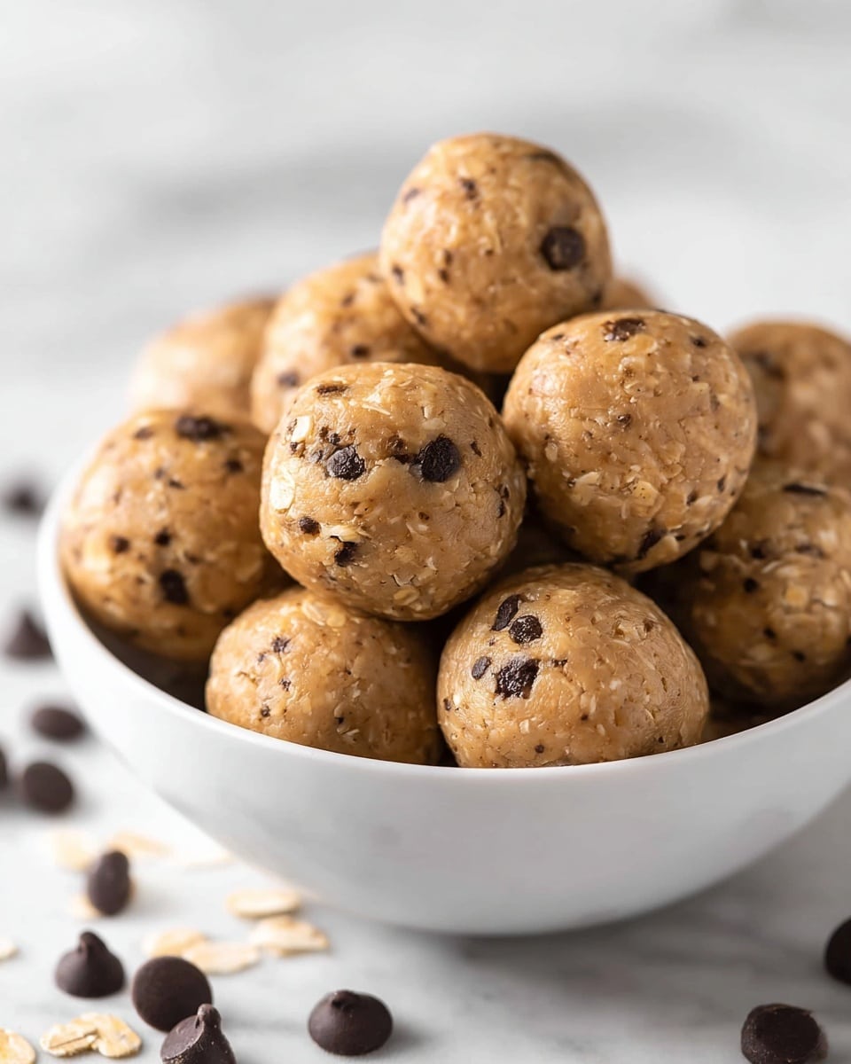 A white bowl filled with about 12 round energy balls that are light brown with visible small dark chocolate chips and bits of oats or nuts mixed inside, giving the balls a slightly textured surface. The balls are piled on top of each other and look soft and chewy. In the background, scattered dark chocolate chips are slightly blurred on a white marbled surface. Photo taken with an iphone --ar 4:5 --v 7