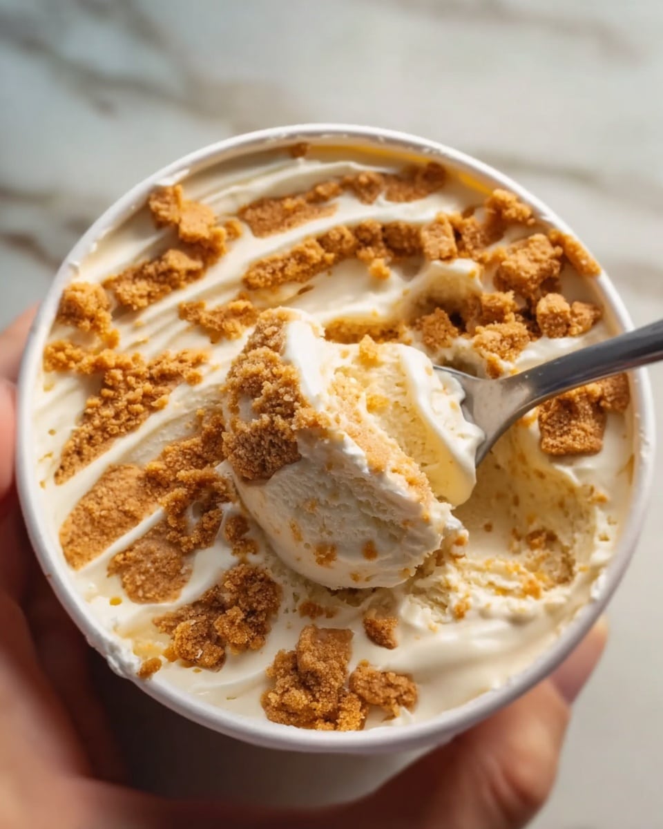 A close-up view of a white round container filled with creamy light beige ice cream layered with thick, crumbly golden-brown cookie pieces swirled throughout, showing a mix of smooth and crumbly textures. A silver spoon is scooping a portion from the bottom right, lifting a blend of the creamy ice cream and chunks of cookie. The container sits against a white marbled surface, with a woman's hand holding it gently on the right side. The layered combination of cream and cookie creates a swirled, textured pattern on the ice cream surface. photo taken with an iphone --ar 4:5 --v 7