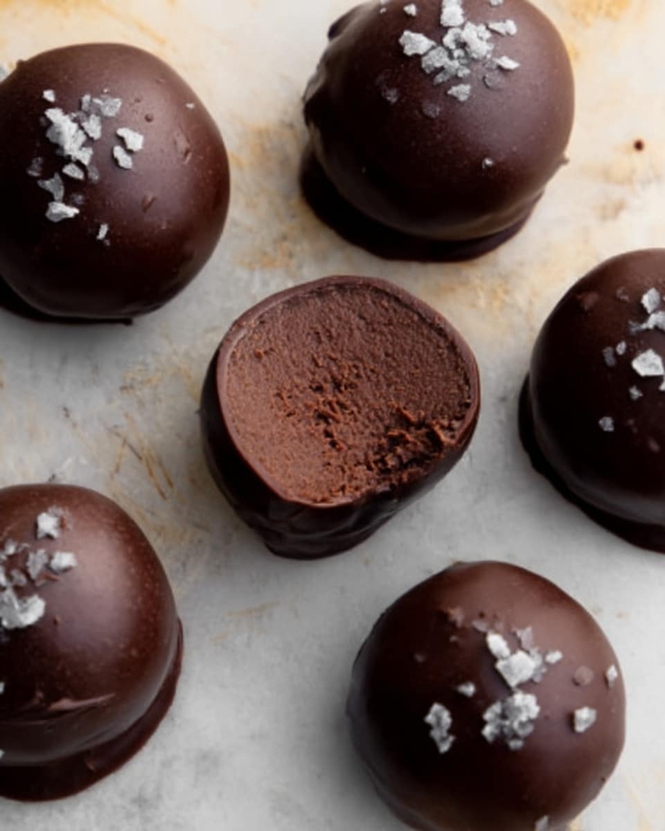 The image shows six round chocolate truffles on a white marbled surface. Five truffles are whole, smooth, and shiny with a dark chocolate coating, and one is cut in half, revealing a dense, rich, dark brown chocolate filling inside. Some of the whole truffles have small white flakes sprinkled on top, adding texture and contrast. The lighting highlights the glossy texture of the chocolate, making the truffles look fresh and delicious. photo taken with an iphone --ar 4:5 --v 7