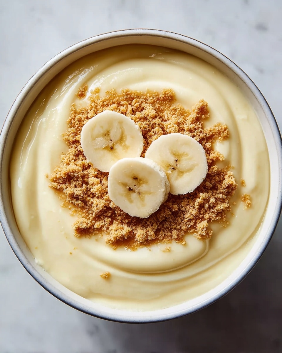 A white bowl filled with smooth, creamy light yellow pudding swirled with soft, rounded ridges. On top, there is a sprinkling of light brown crumbled cookie pieces creating a textured layer. At the center, three round banana slices with pale yellow flesh and light brown spots sit neatly on the crumbs, adding a fresh fruit element. The bowl sits on a white marbled surface. photo taken with an iphone --ar 4:5 --v 7