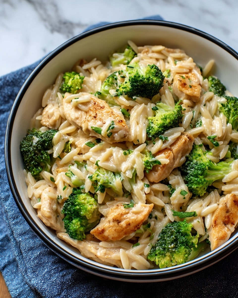 The dish shows one bowl filled with a creamy pasta mix. The bottom layer is light cream sauce covering the base. On top of that, there are light golden-brown cooked chicken pieces scattered around. Bright green broccoli florets are mixed evenly throughout with some pieces tucked under the chicken and pasta. The pasta itself is small and rice-shaped, pale cream in color, and fills the spaces between the other ingredients. The bowl is white with a black rim, placed on a blue cloth, set on a white marbled surface. photo taken with an iphone --ar 4:5 --v 7