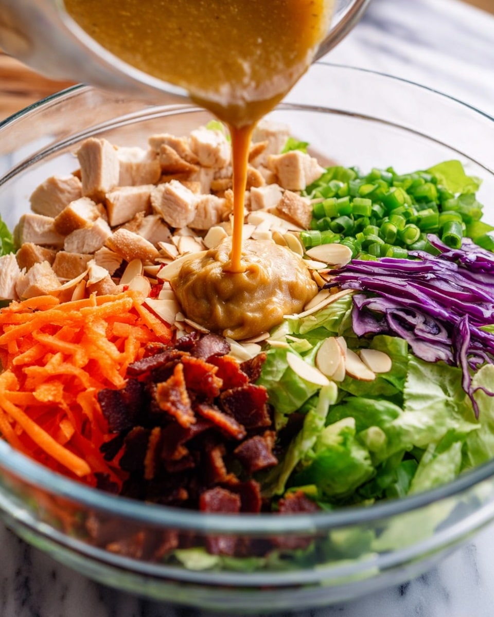 A white plate with black trim holds a colorful layered salad. The base layer is fresh green lettuce mixed with shredded purple cabbage and orange carrots, giving a mix of green, purple, and orange colors with a crisp texture. On top, there are pieces of grilled chicken that look lightly browned and tender. Scattered over the chicken and veggies are sliced light brown almonds, green chopped scallions, and small white sesame seeds. A silver fork rests on the right of the plate, and the whole scene is set on a white marbled texture with pieces of ginger and garlic around, plus a grey cloth to the left. Photo taken with an iphone --ar 4:5 --v 7