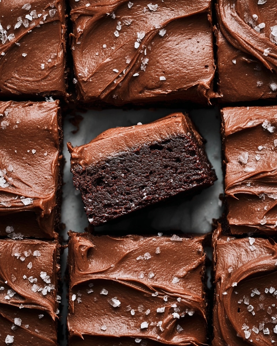 A close-up view of a batch of chocolate brownies cut into rectangular pieces, arranged closely together on a white marbled surface. Each brownie has two layers: a rich dark brown, slightly crumbly chocolate base with visible texture, topped with a thick, smooth spread of glossy milk chocolate frosting that has gentle swirls and peaks. Some brownies are sprinkled with small crystals of white sea salt, creating contrast. One piece is lifted slightly out of line, showing the thickness and moistness of the chocolate base under the creamy frosting. Photo taken with an iphone --ar 4:5 --v 7