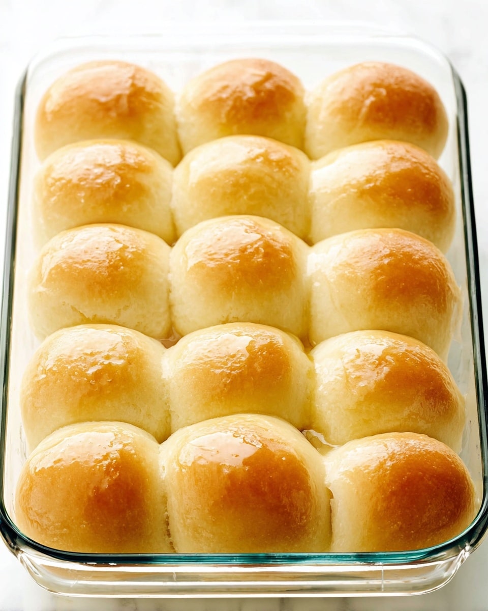 A glass baking dish holds 15 soft, golden-brown dinner rolls arranged in a 3 by 5 grid, each roll having a smooth, shiny top with a light glaze that gives them a slightly glossy look. The rolls are touching each other, showing a uniform, fluffy texture with subtle browning on the tops, while the sides remain a light beige. The dish is placed on a white marbled surface that provides a clean and bright background. photo taken with an iphone --ar 4:5 --v 7